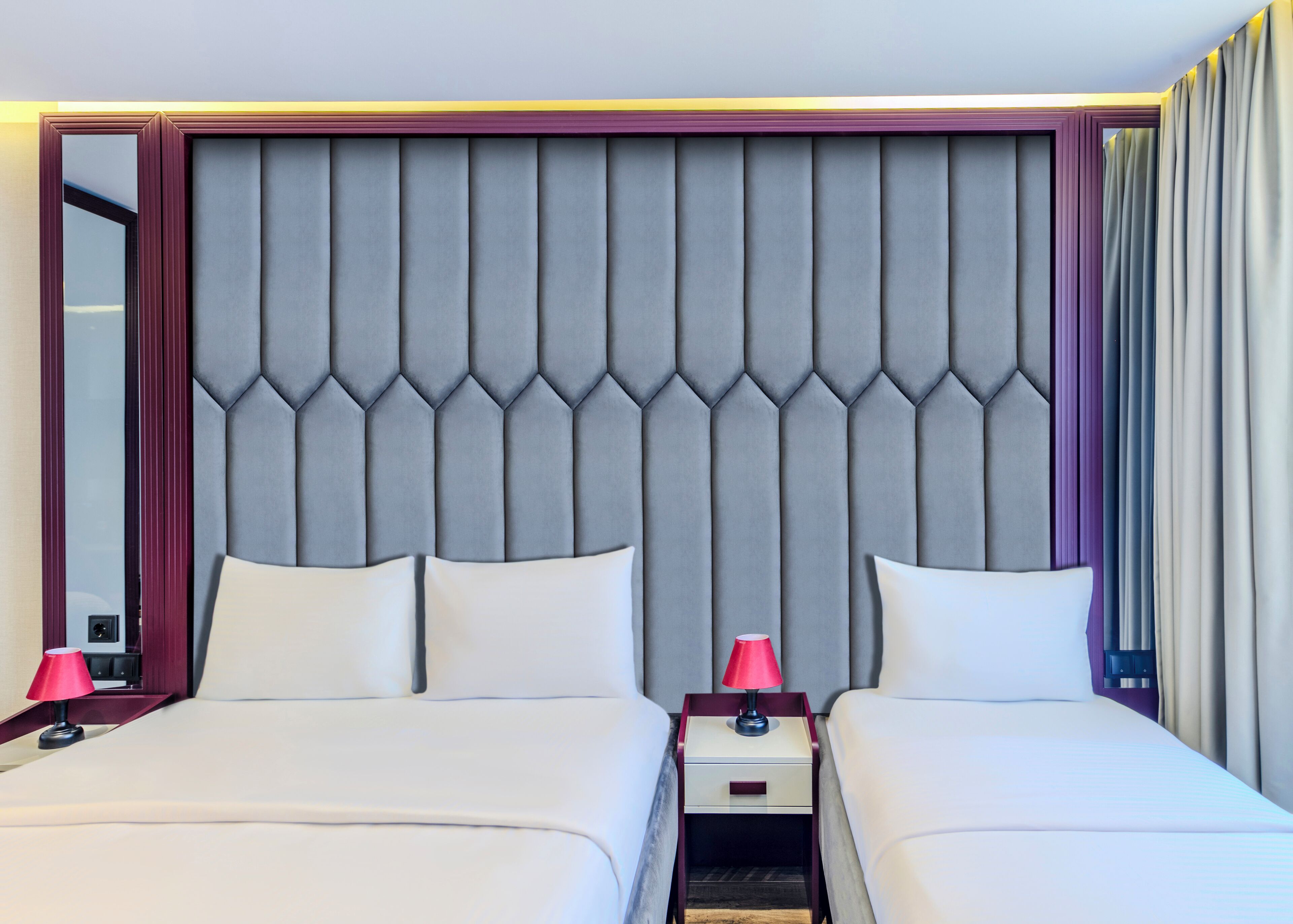 A hotel room with two beds, a headboard with a geometric pattern, a nightstand with a lamp, and a mirror.