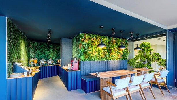Un área de comedor moderna con una paleta de colores azul y verde, que presenta una larga barra con bufé y un espacio de estar con mesas y sillas de madera.