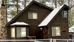 A two-story wooden cabin with a stone chimney, a fenced front yard, and a small porch. - Knotty Pine Tavern-BBC (Big Bear)