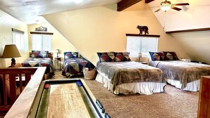 A cozy room with a shuffleboard table, two beds with patterned bedding, a ceiling fan, and a small table with a lamp. - Knotty Pine Tavern-BBC (Big Bear)