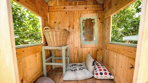 A cozy wooden cabin room with a wicker basket on a stool, a cushion with text, and a polka dot pillow.