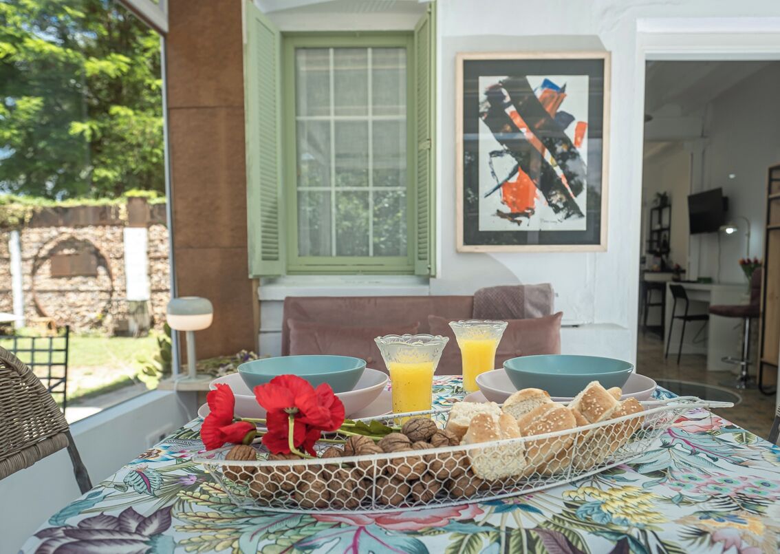A breakfast table with a floral tablecloth, two bowls, two glasses of orange juice, a basket of bread, and a red flower.