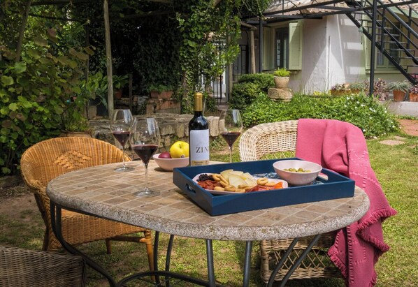 Una mesa redonda con una silla de mimbre, una botella de vino y una caja de aperitivos.