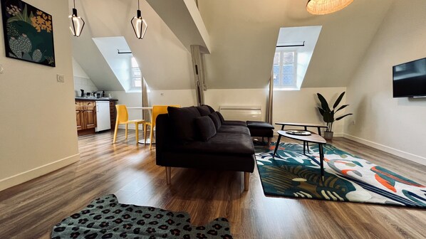 Ein modernes Wohnzimmer mit einem schwarzen Sofa, gelben Sesseln, einem Couchtisch und einem gemusterten Teppich. Im Hintergrund befindet sich eine Küche mit Holzschränken und einem Esstisch.