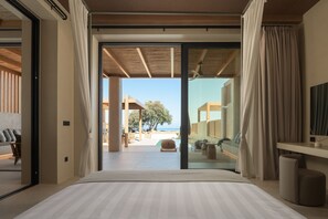A bedroom with a large bed, sliding glass doors leading to a balcony with a view of the beach and a tree. - Cicada Alas Suite, with Private Pool, Gym, BBQ,  Beach & 5*Hotel Access
 (Heraklion)