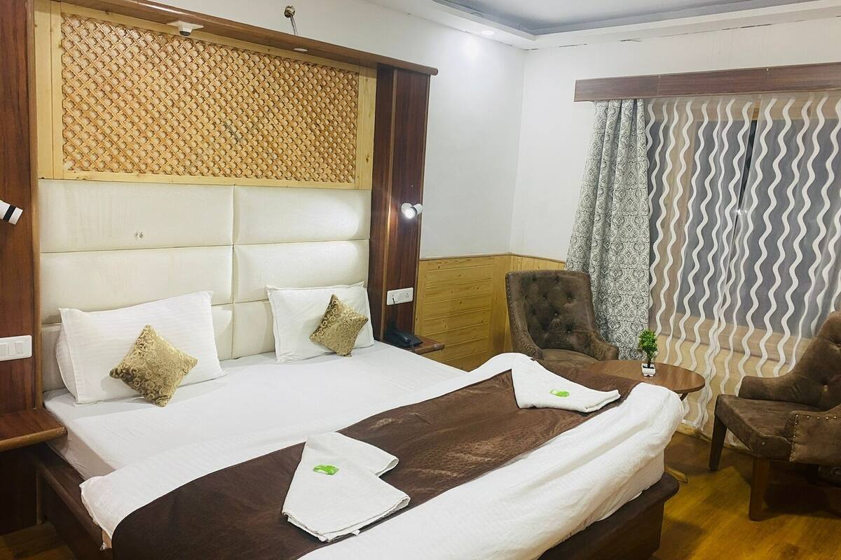 A hotel room with a large bed, a wooden headboard, a brown armchair, a small table with a plant, and a curtain with a wavy pattern.