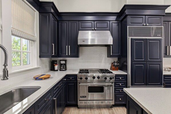 A kitchen with dark cabinets, stainless steel appliances, a window with blinds, and a wooden cutting board.
