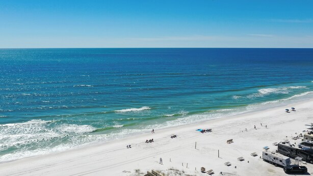 A beach with white sand, clear blue water, and a few people and vehicles.