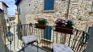 Ein Balkon mit gepolstertem Stuhl, zwei Blumenkästen und einem Steinhaus mit blauen Fensterläden.