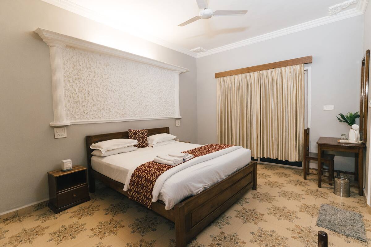 Quarto com cama, mesa de cabeceira, janela com cortinas e ventoinha no teto.