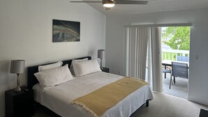 A bedroom with a bed, a ceiling fan, a painting, and a view of the outdoors.