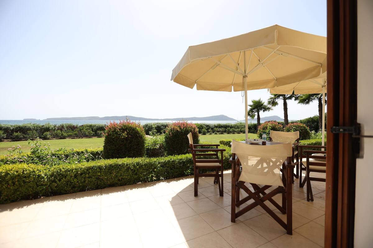 An outdoor patio with a table and chairs under a large umbrella, overlooking a landscaped garden and distant hills.