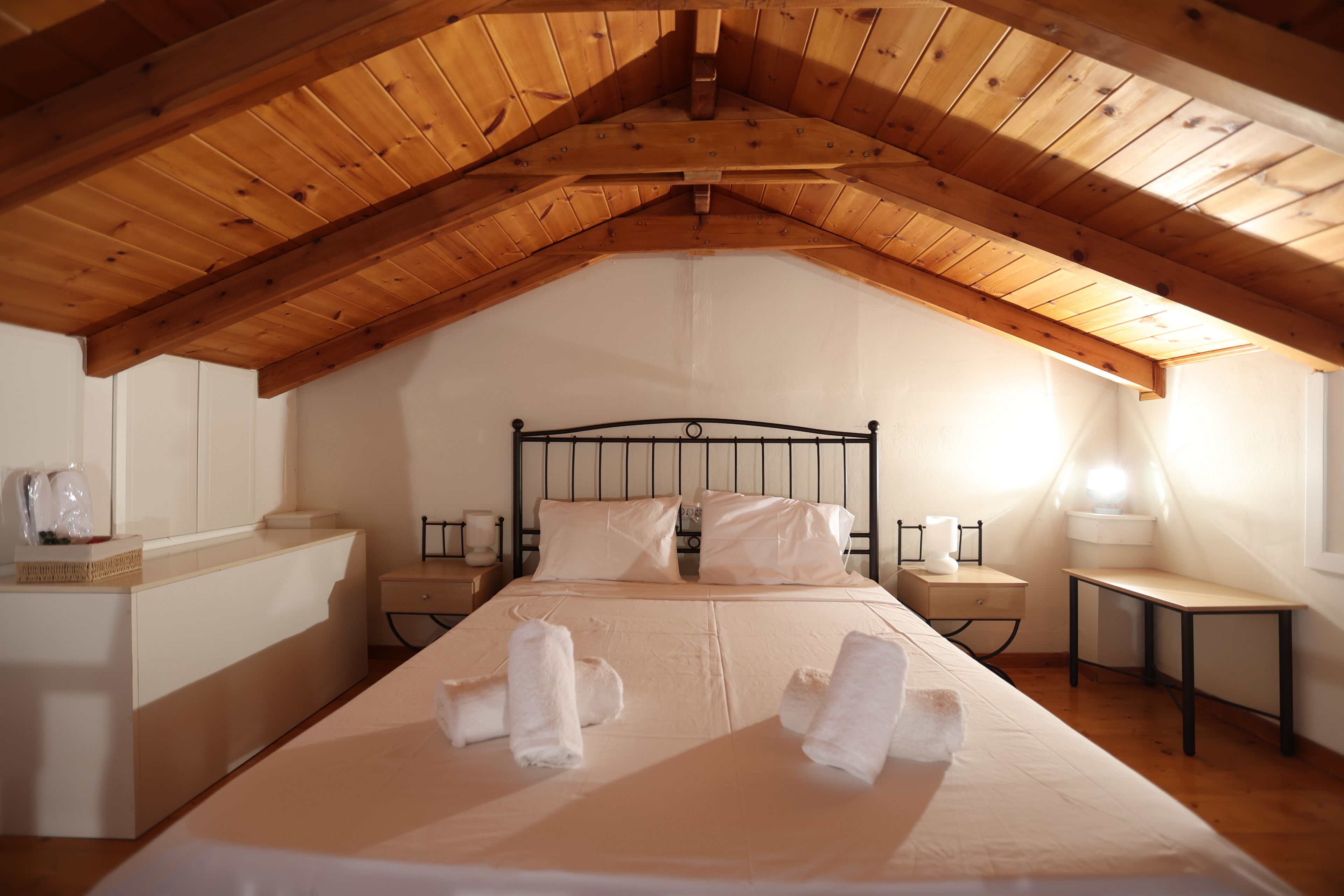 A bedroom with a wooden ceiling, a bed with white linens, two bedside tables, and a nightstand with a lamp.