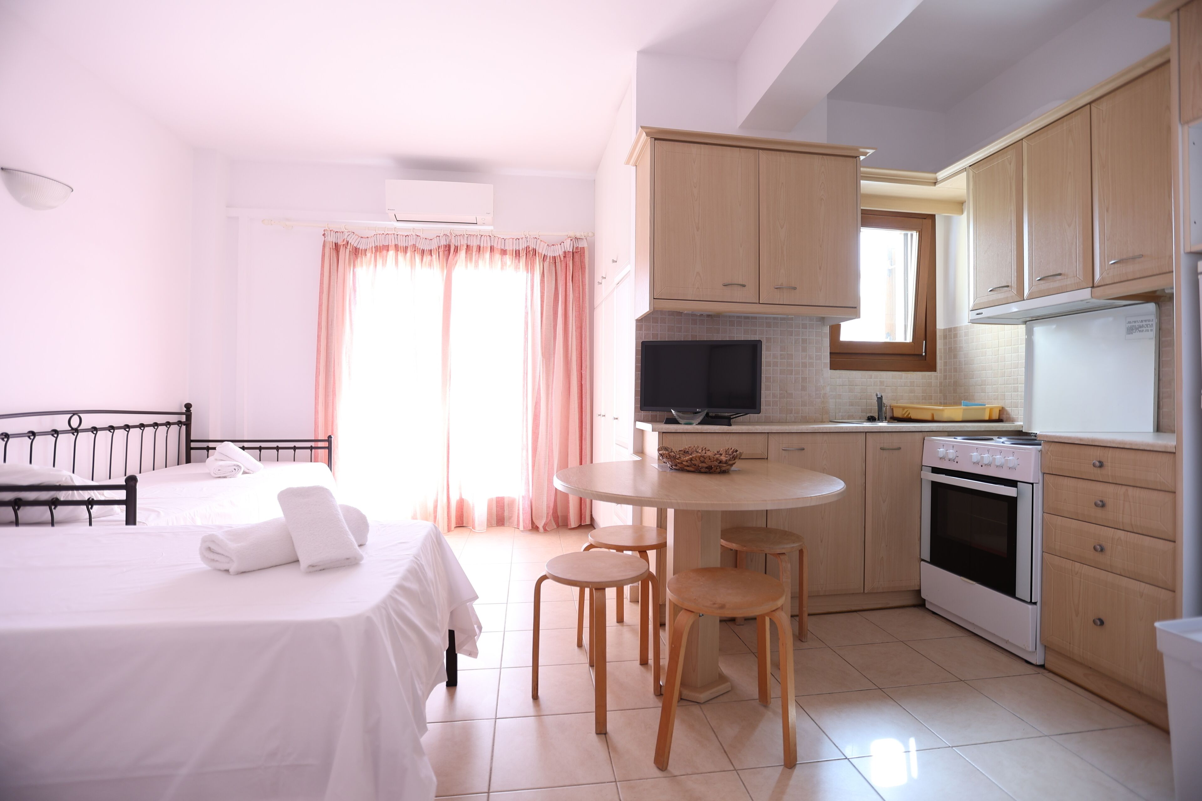 Une chambre d’hôtel comprenant un lit, une petite table à manger, un coin cuisine avec des placards en bois, une cuisinière et un téléviseur.