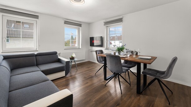 A modern living room with a grey sectional sofa, a wooden dining table with four chairs, and a flat-screen TV mounted on the wall. - ADN Apartments, modern and minimalist (Koetz)