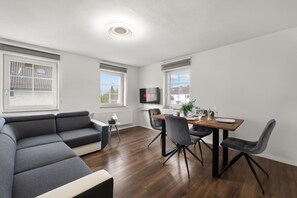 A modern living room with a grey sectional sofa, a wooden dining table with four chairs, and a flat-screen TV mounted on the wall. - ADN Apartments, modern and minimalist (Koetz)