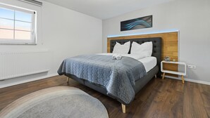A modern bedroom with a large bed, a nightstand, a wall-mounted light, and a window with blinds. - ADN Apartments, modern and minimalist (Koetz)