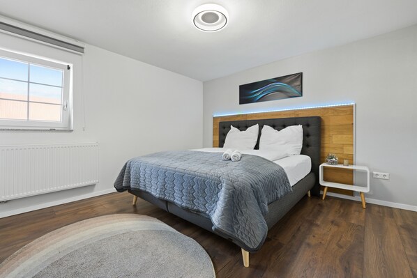 A modern bedroom with a large bed, a nightstand, a wall-mounted light, and a window with blinds. - ADN Apartments, modern and minimalist (Koetz)