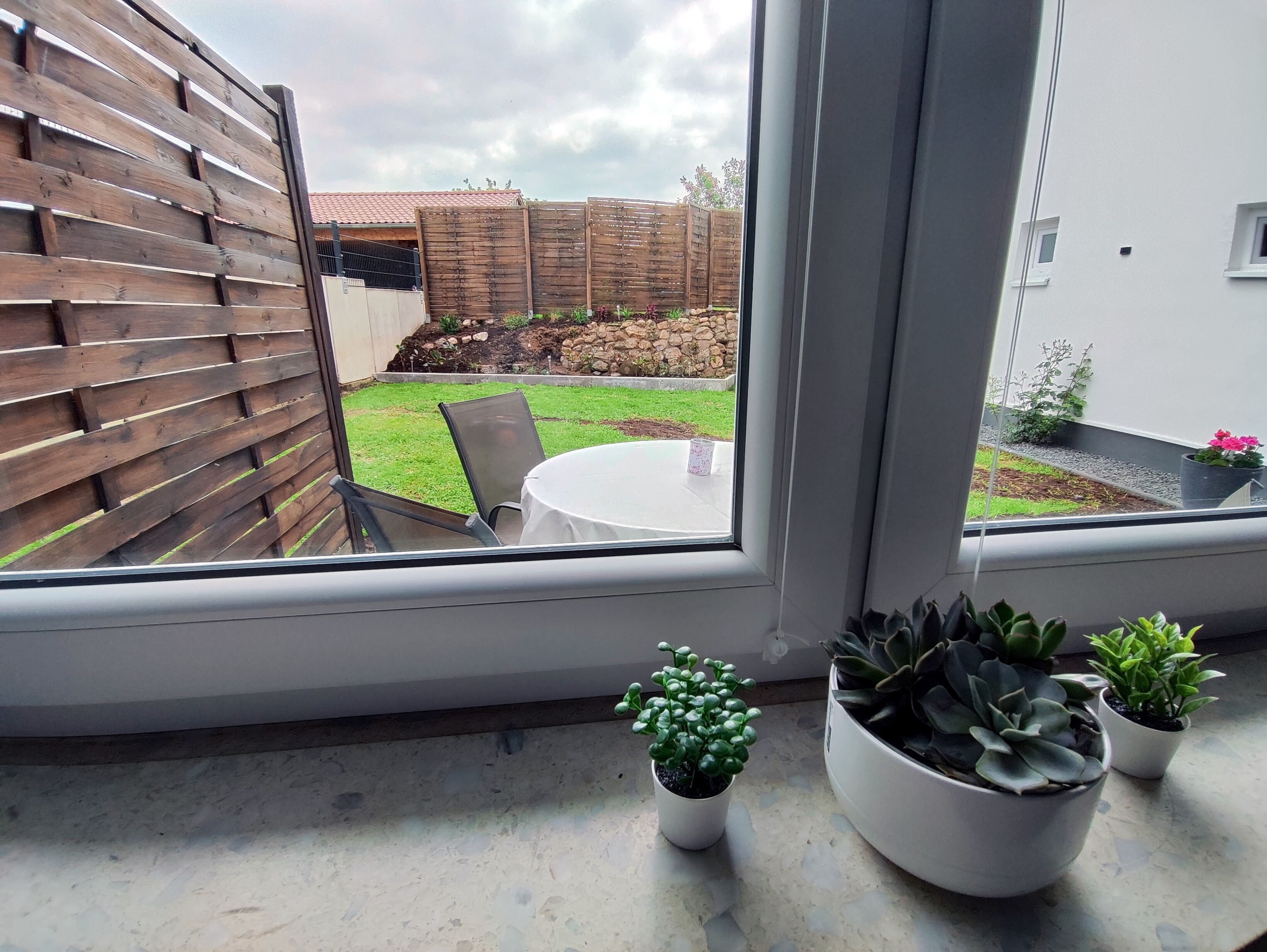 Vista desde una ventana a un patio con mesa y sillas, una cerca de madera y plantas en macetas en el alféizar.