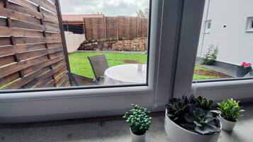 Vista desde una ventana a un patio con mesa y sillas, una cerca de madera y plantas en macetas en el alféizar.