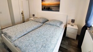 A bedroom with a bed, two bedside tables, a nightstand, a wardrobe, and a painting of a sunset over the sea. - Vacation apartment Heide I (Walsrode)