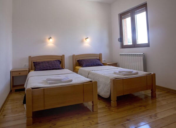 A room with two wooden beds, each with a pillow and a neatly folded towel. There is a nightstand with a lamp on each side of the beds. A window with blinds is visible on the wall. - Forest Edge Two-Bedroom Apartment in Zabljak (Prisoje)