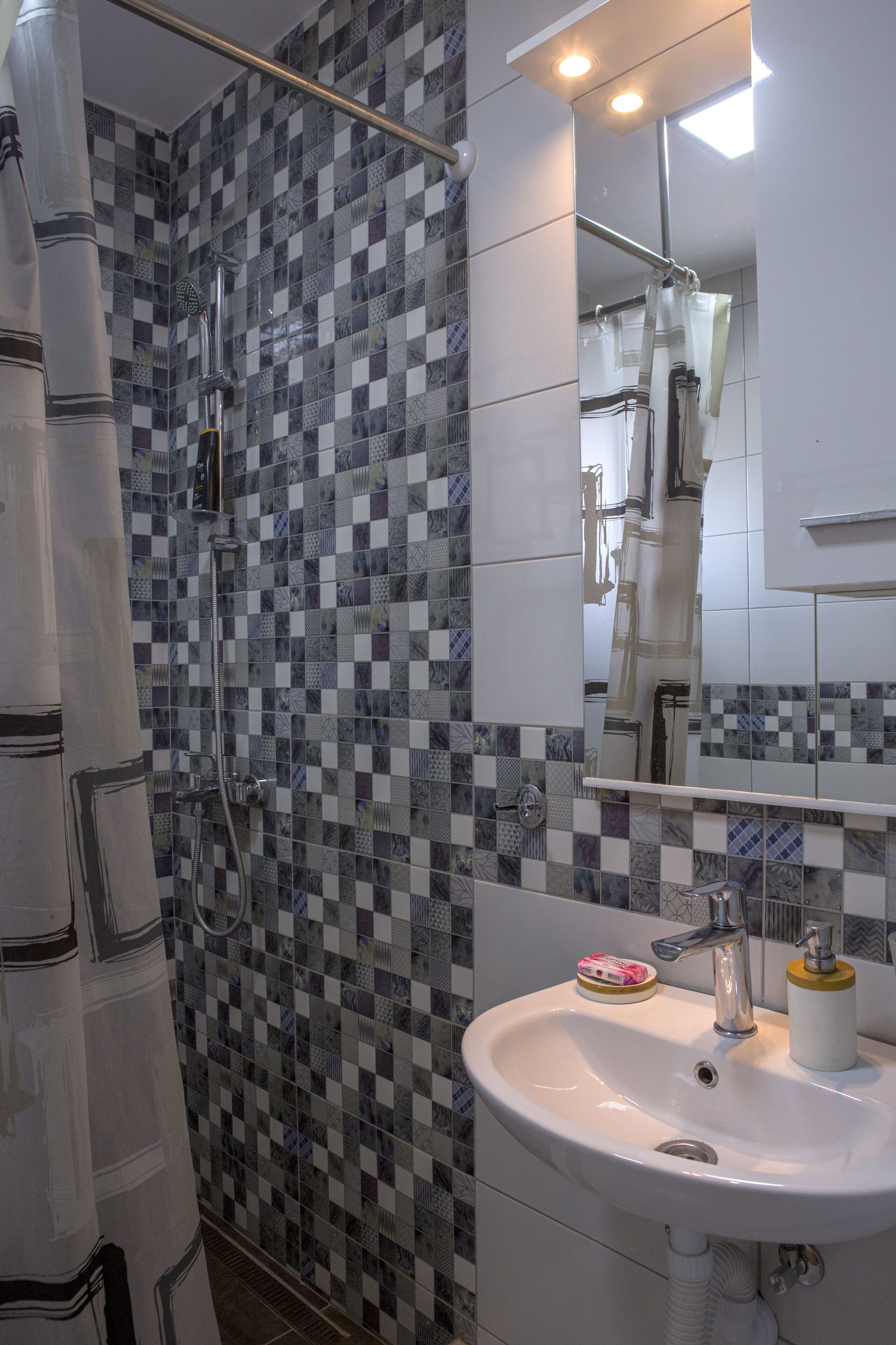 A bathroom with a checkered tile wall, a shower, a sink with a mirror, and a toilet.