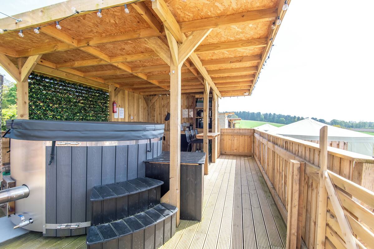 Un espacio al aire libre techado con jacuzzi, terraza de madera y vista a un paisaje verde.