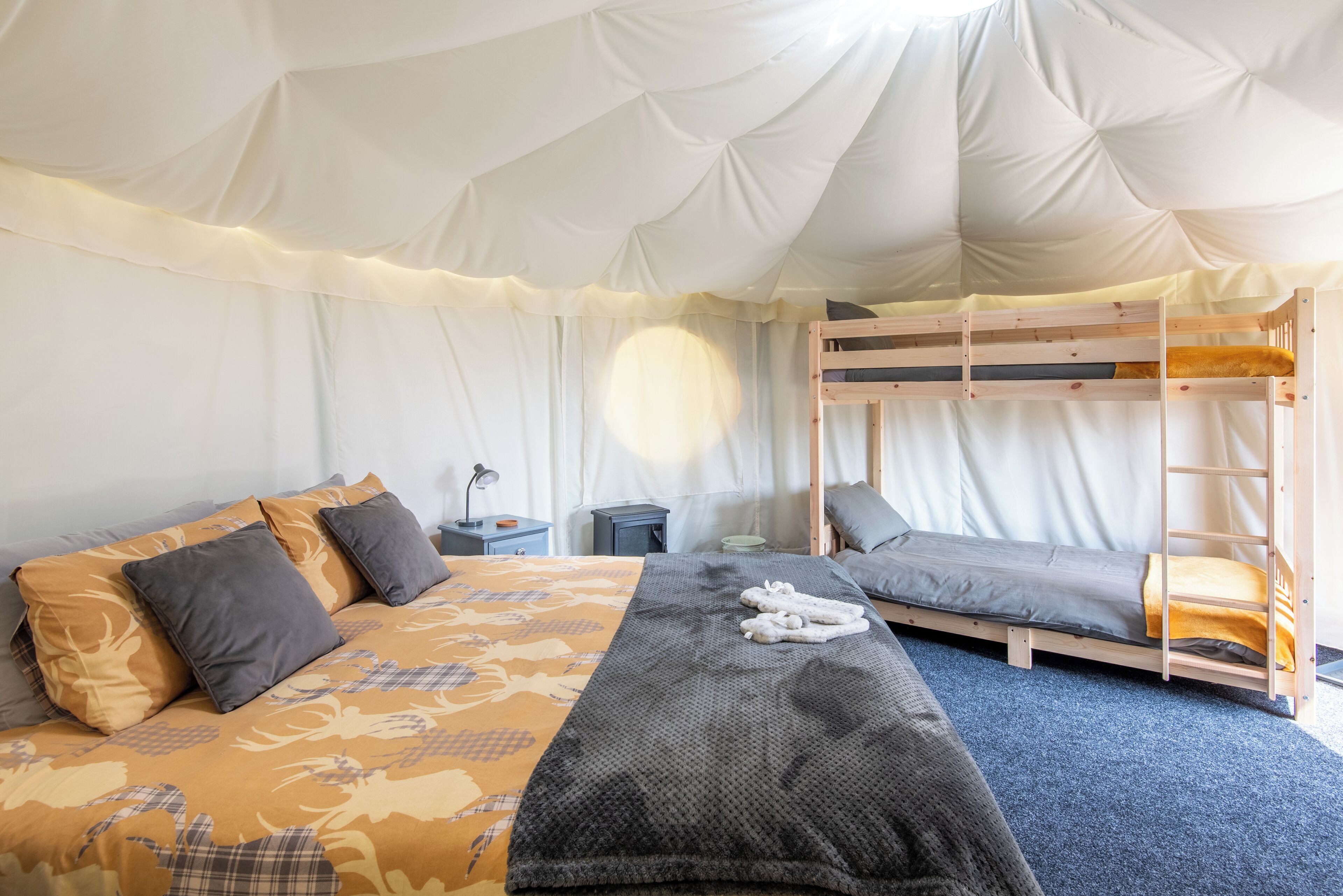 A tented accommodation with a bunk bed, a bed with patterned bedding, and a bedside table with a lamp.