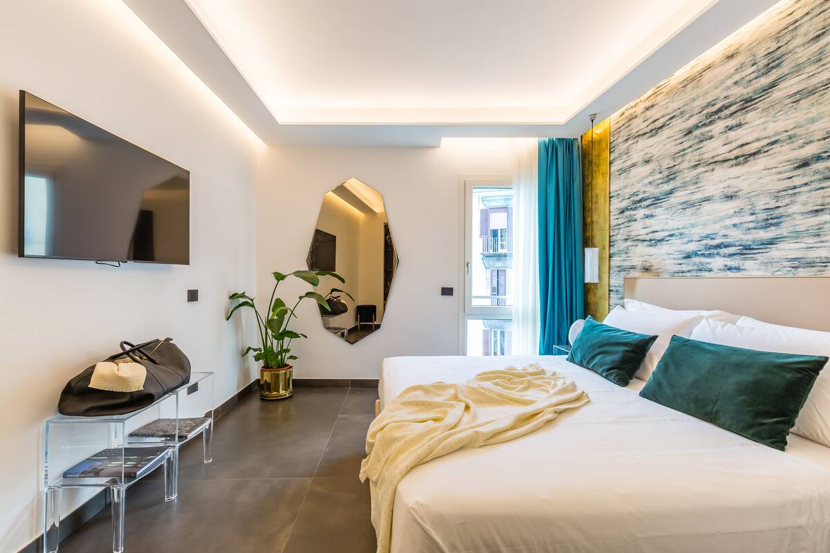 Habitación de hotel moderna con una cama grande, un televisor de pantalla plana montado en la pared, una planta en maceta y un banco transparente con un bolso encima.