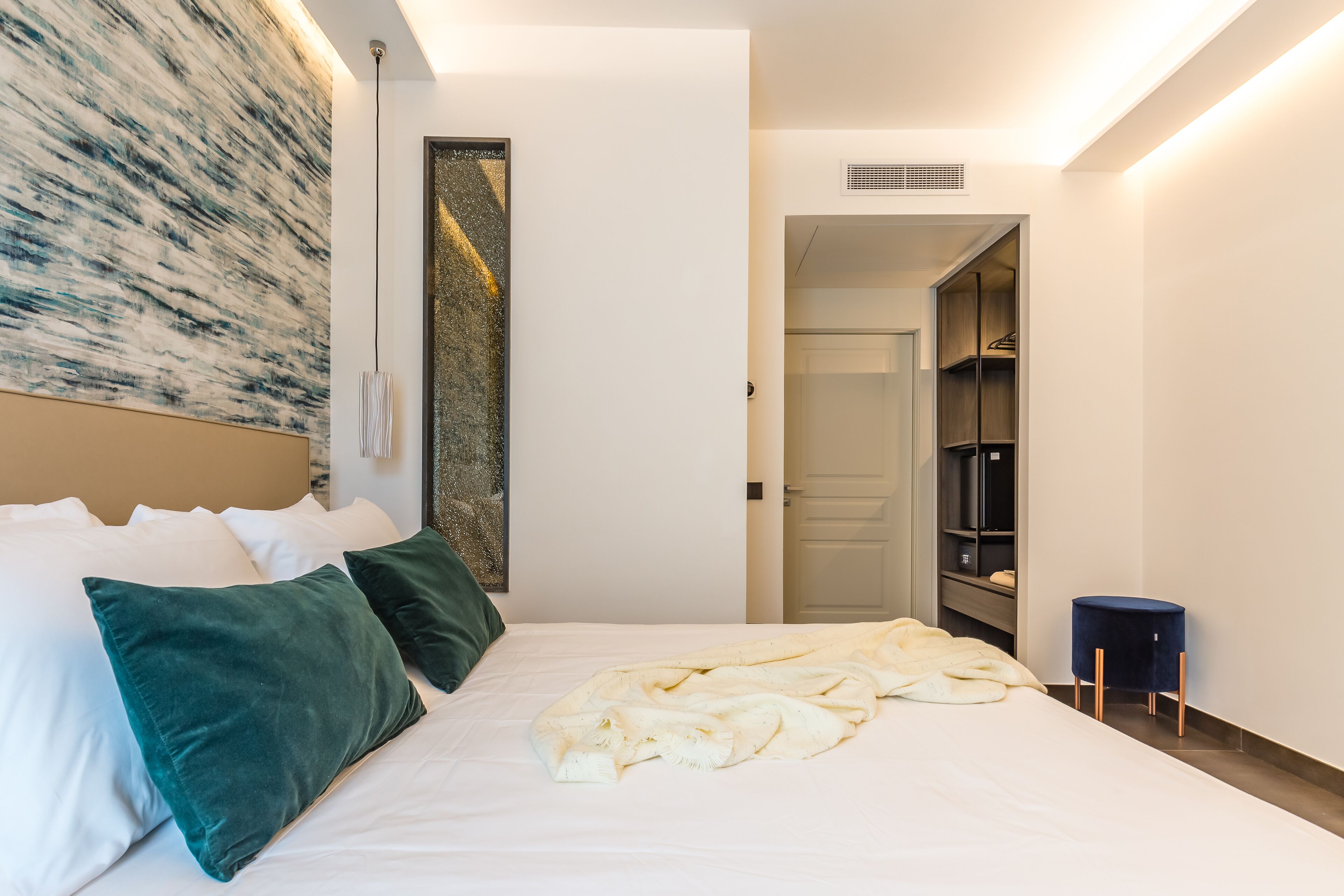 A modern hotel room with a neatly made bed, a large abstract painting on the wall, and a walk-in closet with shelves.