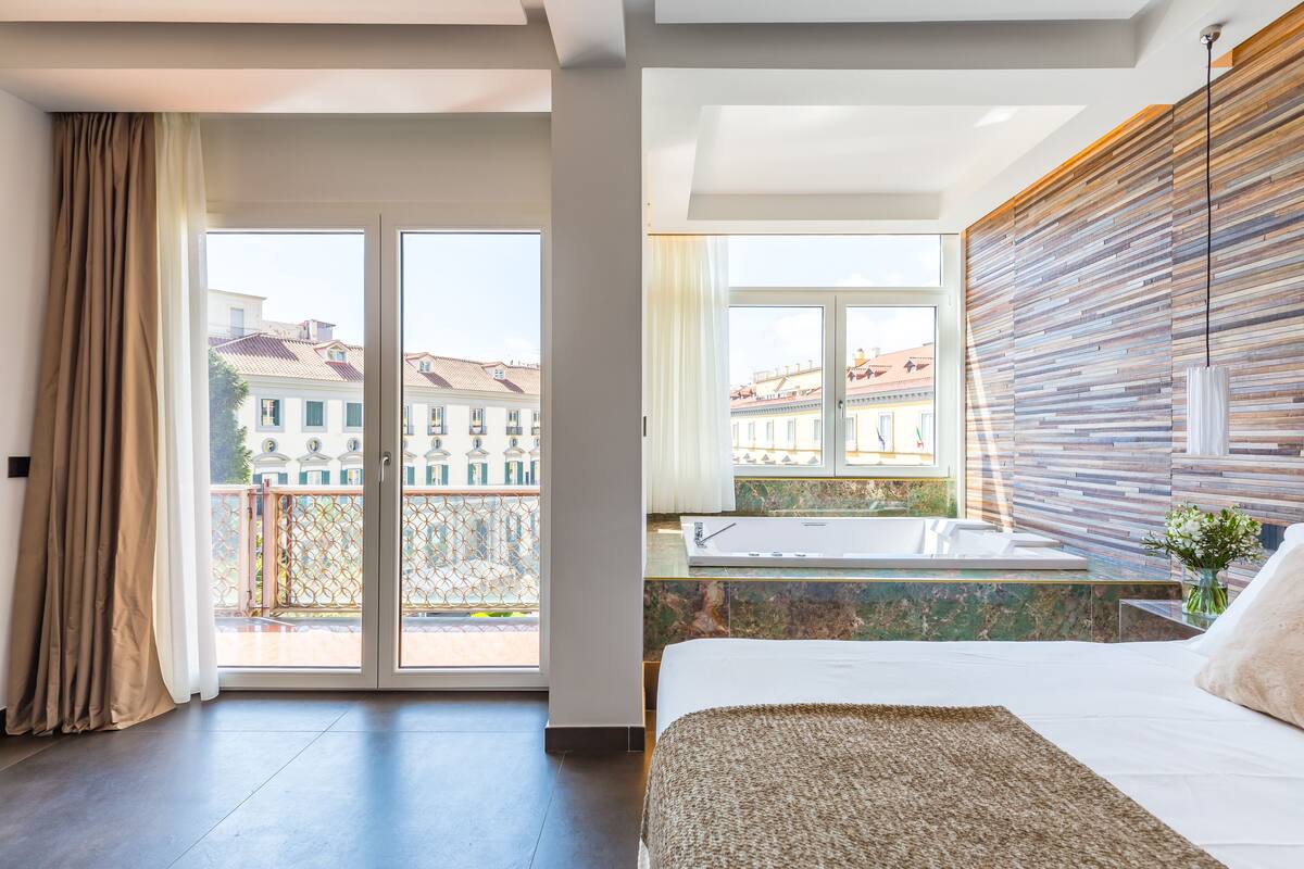 Una habitación de hotel moderna con una cama grande, un balcón con cristales y vistas a edificios.