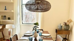 A dining area with a wooden table set for four, a hanging woven light fixture, and a window with curtains. - Eco Luxury apartment Campo de Ourique LISBON center (Lisboa)