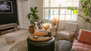 A cozy living room with a round coffee table, a sofa, and a chair. There is a window with blinds, a door, and a wall-mounted artwork of a flamingo.