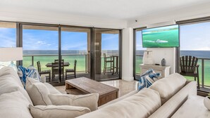A spacious living room with a large sliding glass door leading to a balcony with a view of the ocean. There is a flat-screen TV mounted on the wall, a wooden coffee table, and a beige sofa with blue and white cushions. - Beachfront Wraparound Balcony ~ Golf Cart ~ Pool (Miramar Beach)