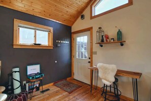 A room with a wooden ceiling, a door with a window, a small table with a white fluffy stool, and a shelf with various items. - The Little Lodge Leadville — 2BR with Hot Tub, Pet-OK, 1.5 Blocks to Harrison (Leadville)