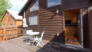 Eine Holzhütte mit einer Terrasse, einem Essbereich mit Stühlen und einer durch eine offene Tür einsehbarer Küche.