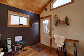 A room with wooden ceiling, a door, a window, a shelf with bottles, a small table, a chair, and a guitar.