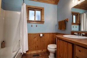 A bathroom with wooden paneling, a white toilet, a sink with a mirror, and a window with a wooden frame. - The Little Lodge Leadville — 2BR with Hot Tub, Pet-OK, 1.5 Blocks to Harrison (Leadville)