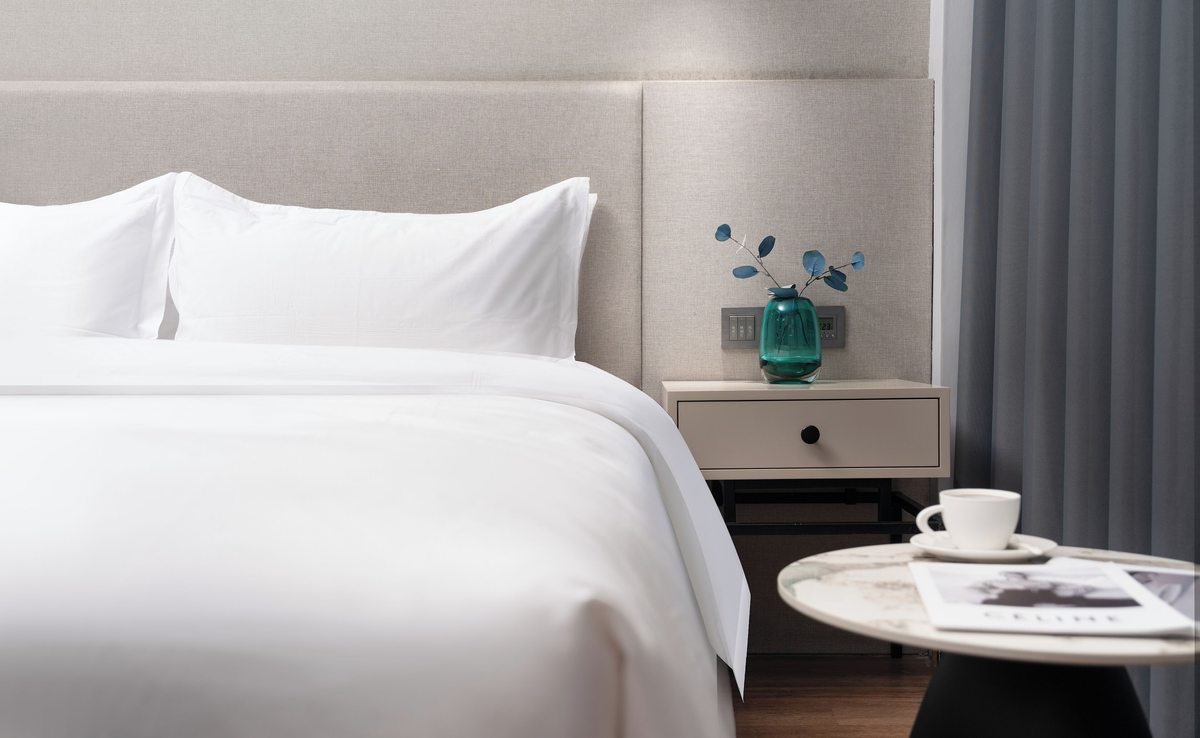 A neatly made bed with white linens, a bedside table with a vase and a clock, and a small round table with a cup and a magazine.