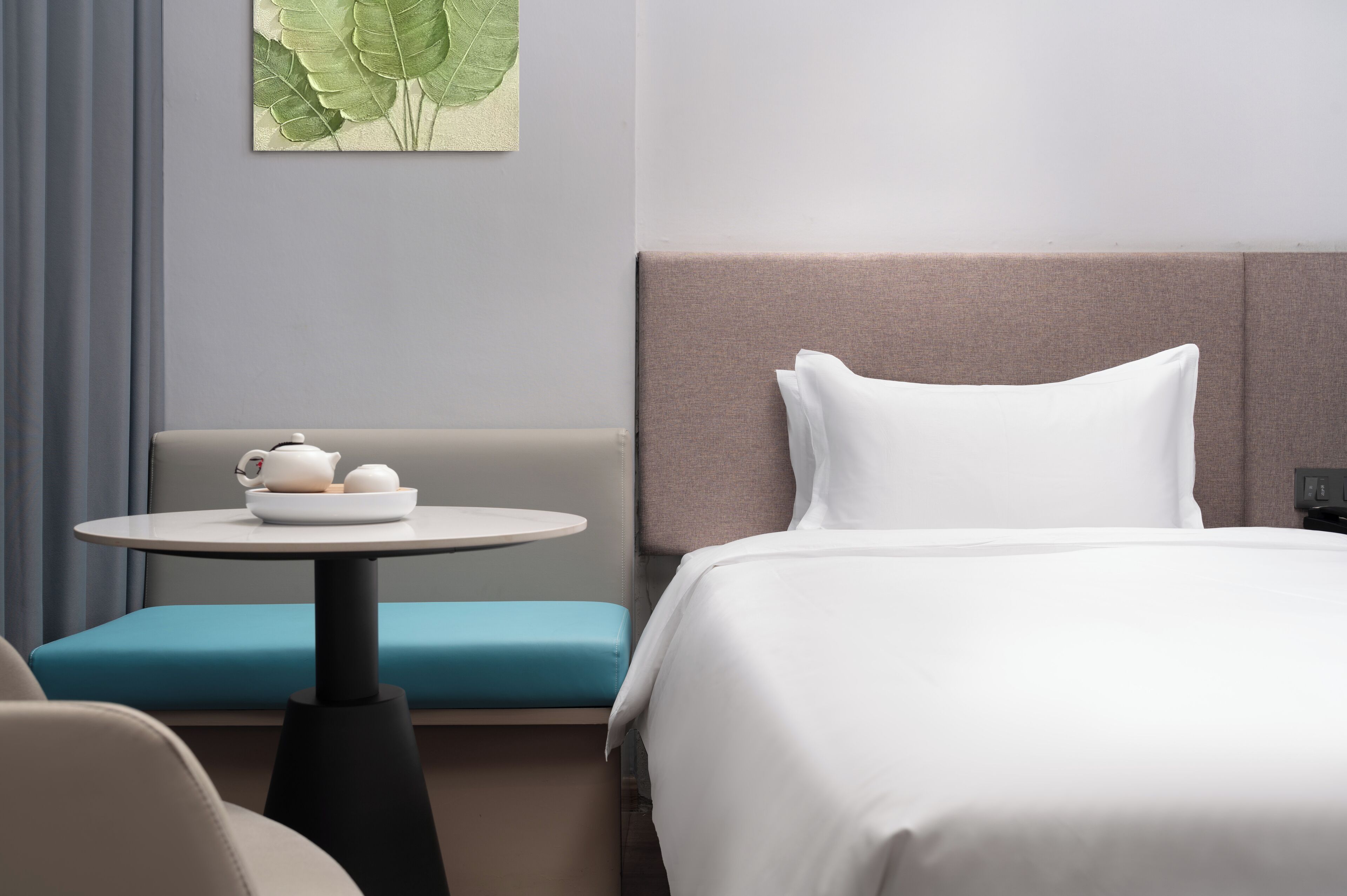 Quarto de hotel com cama, mesa redonda com bule e xícaras, um banco acolchoado azul e uma obra de arte pendurada na parede.