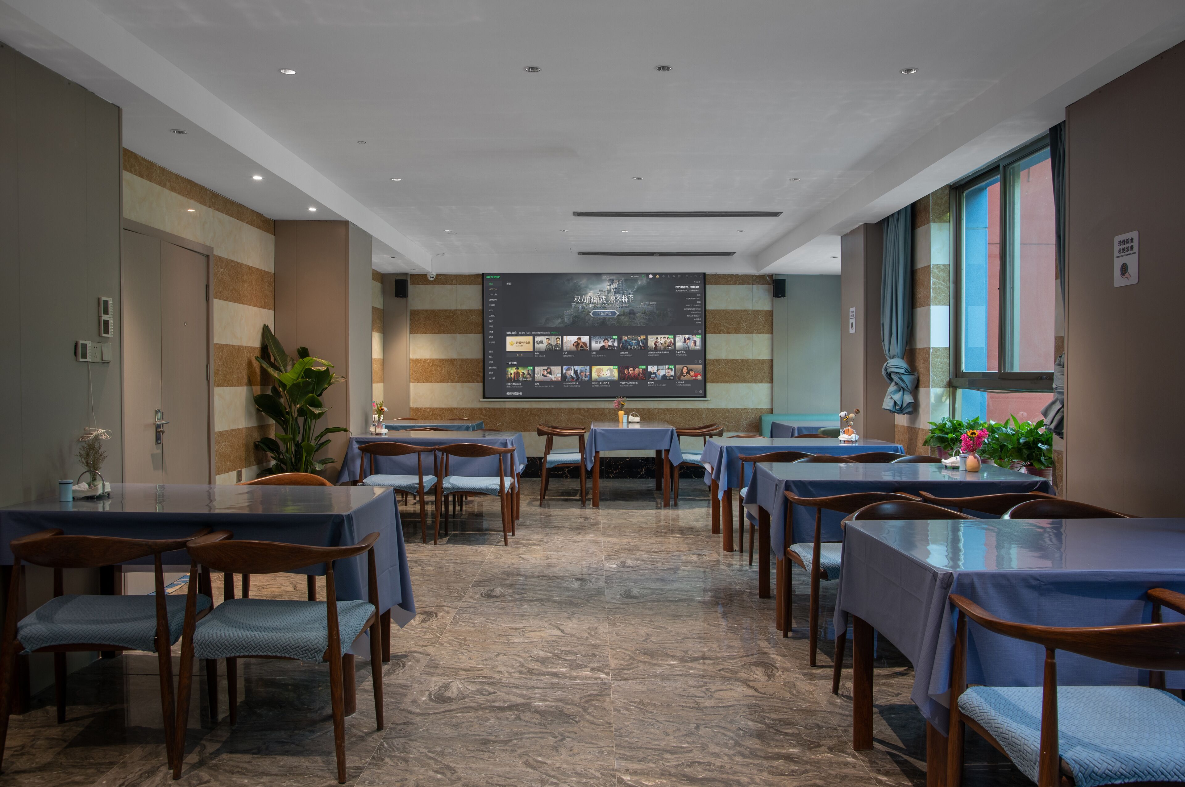 A restaurant with multiple tables and chairs, a large screen displaying menu items, and a potted plant.