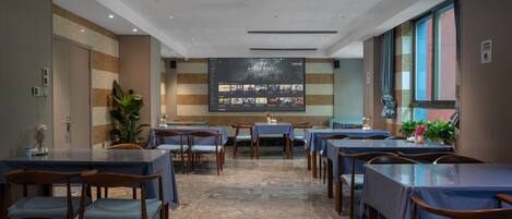 A restaurant with multiple tables and chairs, a large screen displaying menu items, and a potted plant.