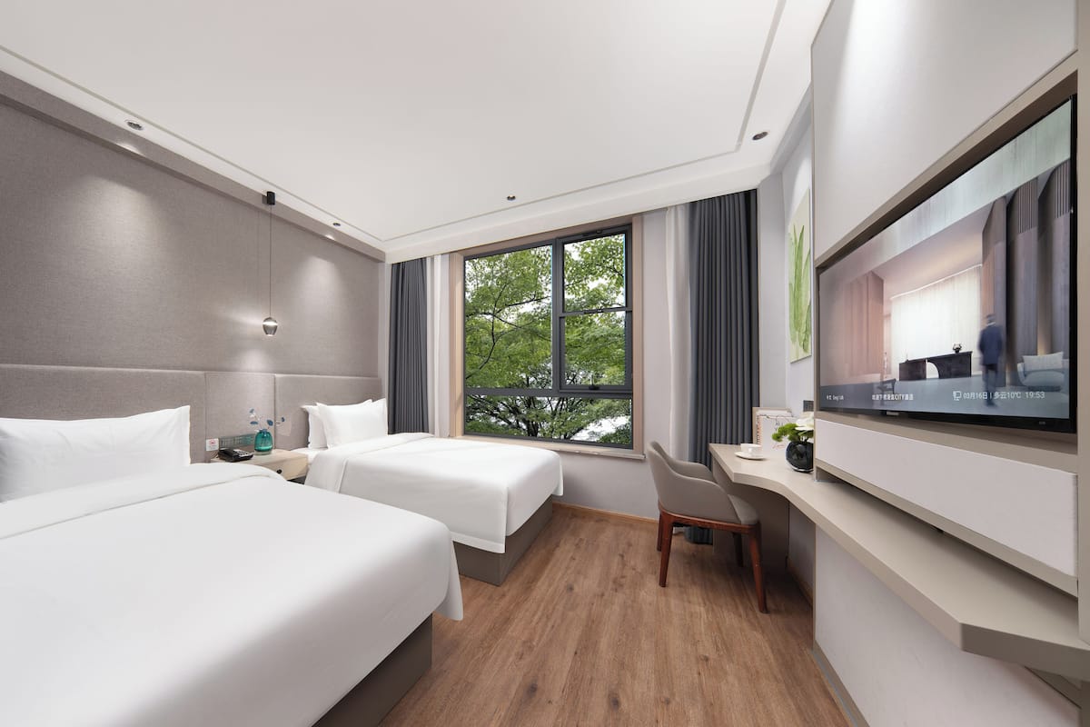 A hotel room with two beds, a desk, a chair, a TV, and a window with a view of trees.