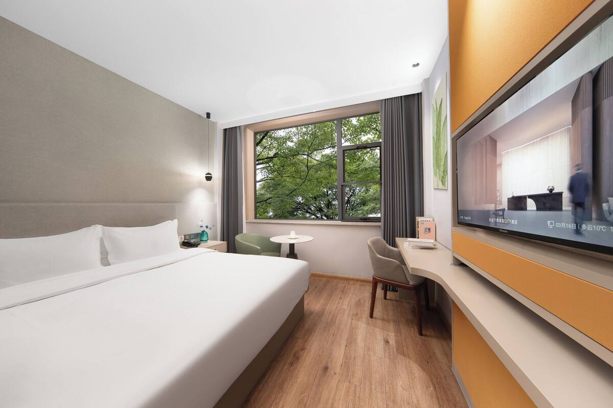 A hotel room with a bed, a desk, a chair, a TV, and a window with a view of trees.