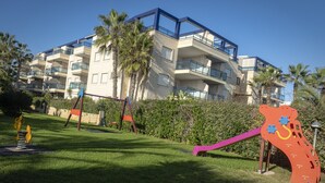 A playground with swings and a slide, surrounded by a well-maintained lawn and palm trees. - DORASAL bl.A bajo Solo Familias! (Xeraco Playa)