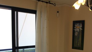 A bedroom with a large window, a bed with floral bedding, a nightstand with a lamp, and a painting on the wall. - DORASAL bl.A bajo Solo Familias! (Xeraco Playa)