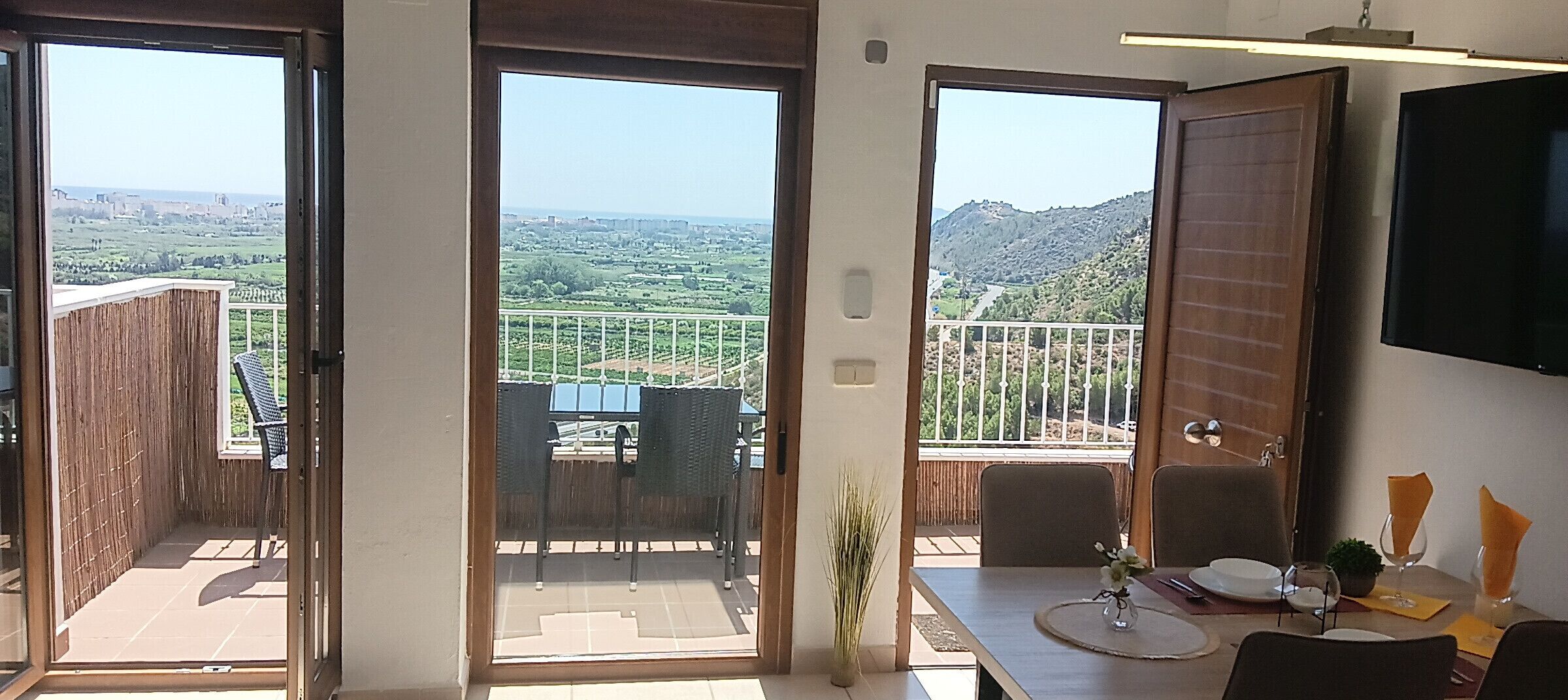 A dining area with a table set for two, a view of a balcony with a railing, and a television mounted on the wall.