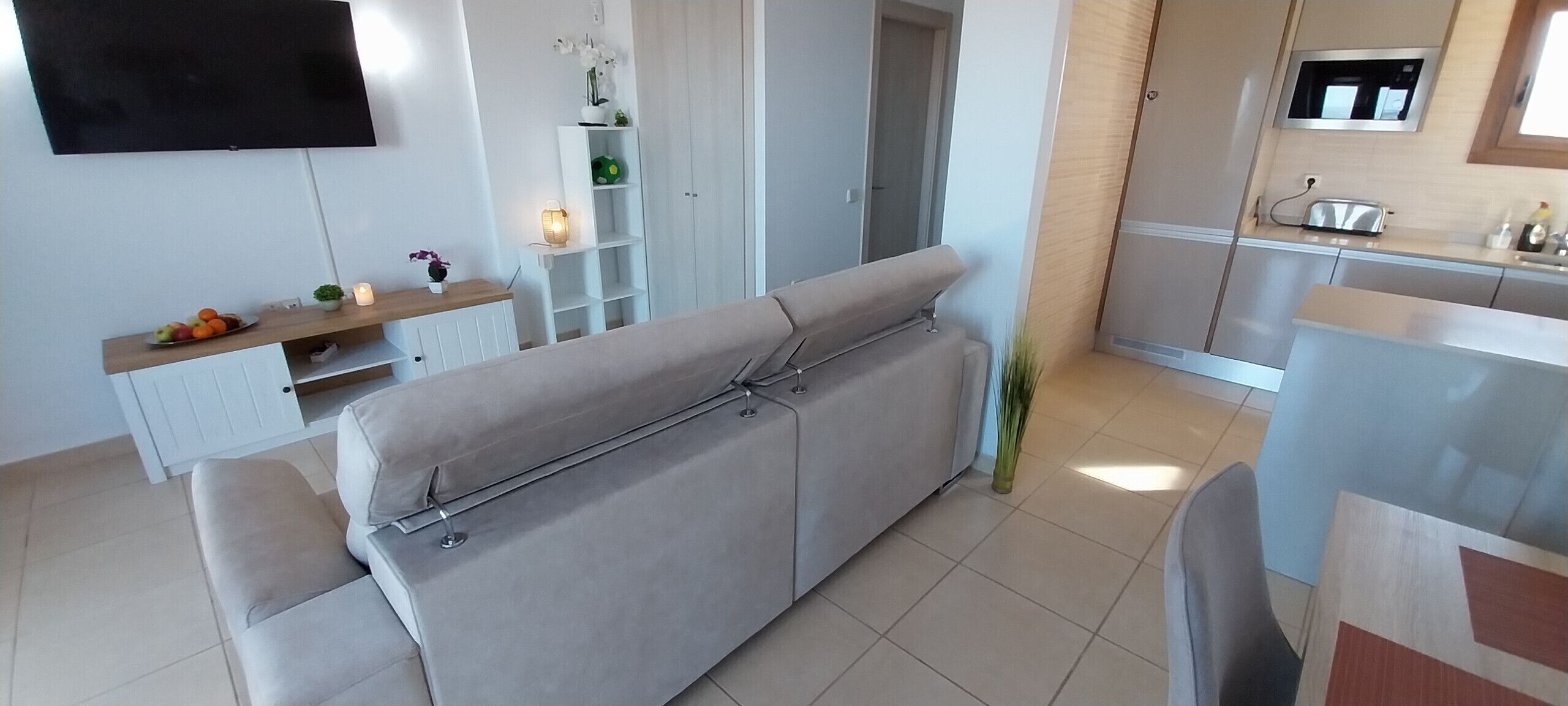 Ein modernes Wohnzimmer mit grauem Sofa, einem an der Wand befestigten Fernseher, einem kleinen Tisch mit Obst und einer Kerze sowie einer Küchenzeile mit Mikrowelle und Toaster.
