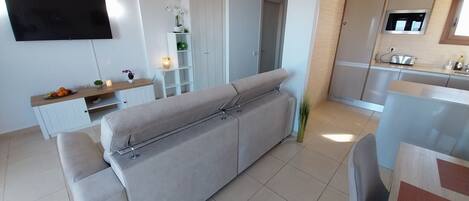 Ein modernes Wohnzimmer mit grauem Sofa, einem an der Wand befestigten Fernseher, einem kleinen Tisch mit Obst und einer Kerze sowie einer Küchenzeile mit Mikrowelle und Toaster.